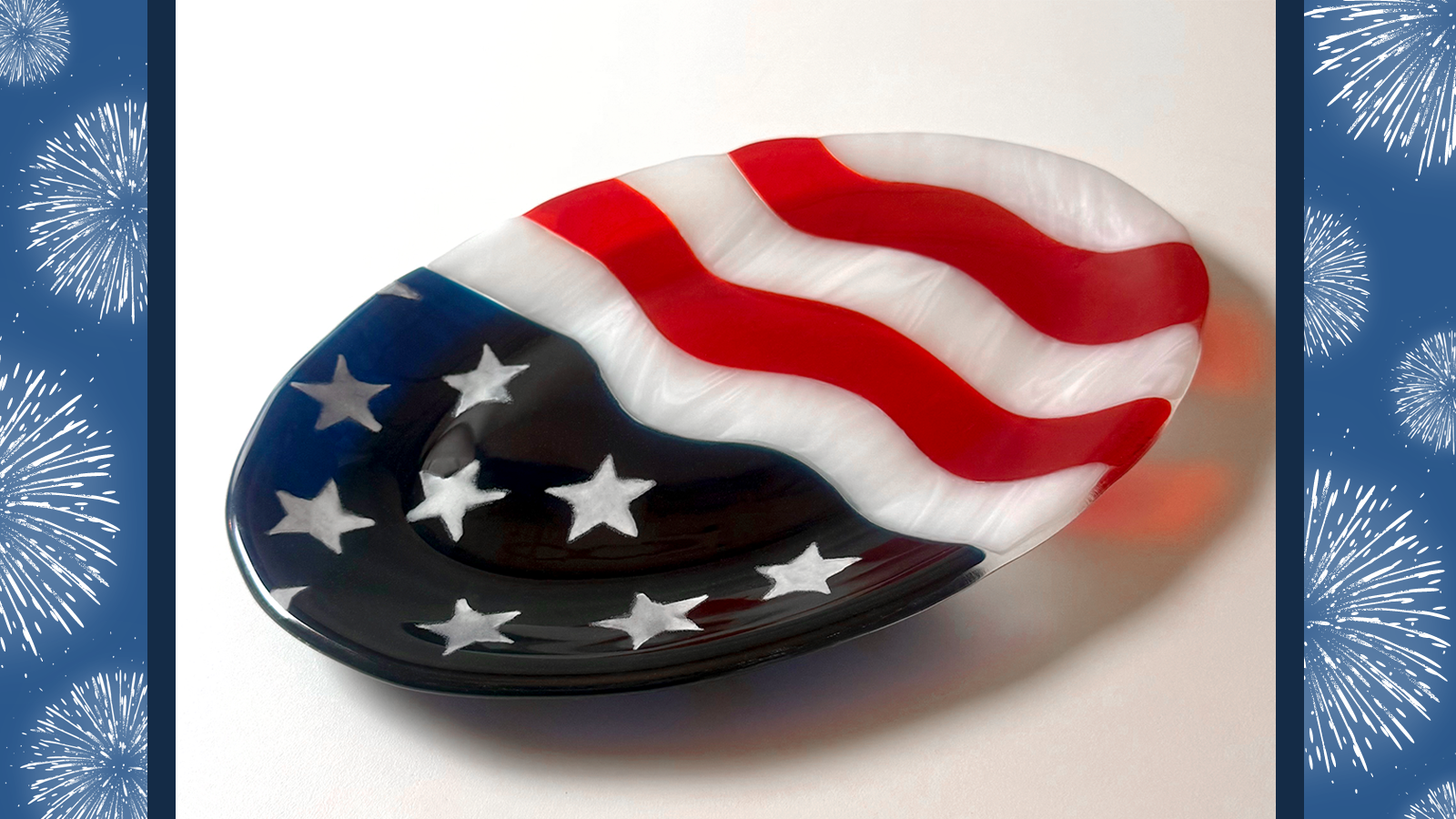 An oval platter made of fused glass on a white background. One half of the platter is dark blue with painted white stars and the other half is wavy bands of red and white, mimicking the American flag.
