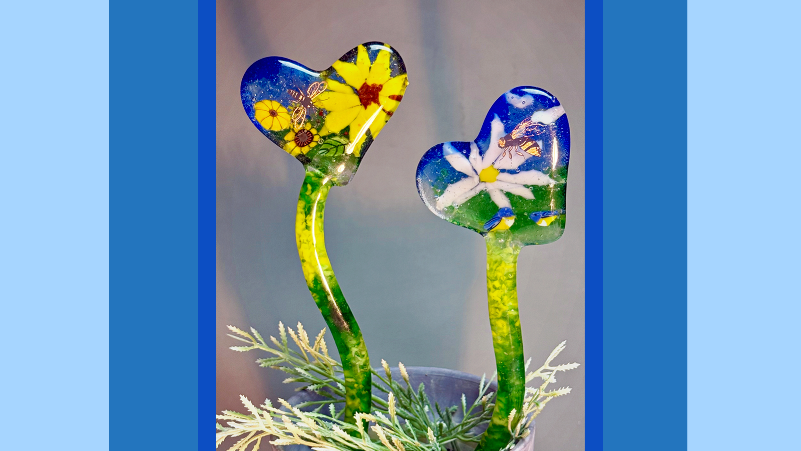 Two stakes made of fused glass. They have curving stems and are each topped with a heart. They are each filled with various flower designs.